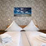 Doppelbett mit weißen Laken, zwei Kissen und gefalteten Handtüchern. Wandbild zeigt Strand und Himmel.