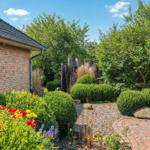 Hinterhof mit gepflastertem Weg, grünen Sträuchern und Blumen vor Backsteinmauer.