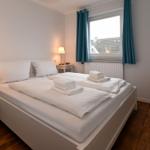 Schlafzimmer mit Doppelbett, zwei Nachttischen und Fenster mit blauen Vorhängen.