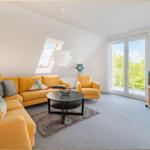 Wohnzimmer mit gelbem Sofa, Rundtisch und TV. Balkon und Dachfenster mit Blick.