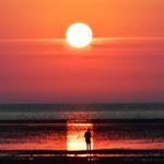 Ein Mensch steht am Strand und blickt auf eine große Sonne, die sich am Horizont befindet.