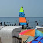 Strand mit weißen Liegestühlen, buntem Sonnenschirm und Segelboot im Wasser.