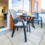 Terrasse mit Tisch und zwei Stühlen. Blick ins Wohnzimmer mit Küche.