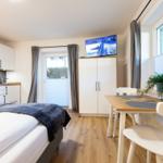 Schlafzimmer mit Küchenzeile, Esstisch und Fernseher. Holzboden und große Fenster.