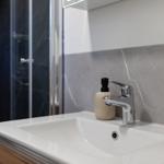 Waschbecken mit Wasserhahn und Seifenspender vor grauem Fliesenspiegel.