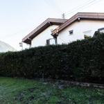 Haus mit weißer Fassade und Holzverkleidung, umgeben von Hecke und Grasfläche mit Bergen im Hintergrund.