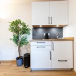 Küche mit weißen Schränken, Holzoberfläche, Spüle und Kaffeemaschine. Pflanze und Stehlampe daneben.