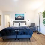 Schlafzimmer mit Bett, Sessel, Tisch und Pflanze. Wandbild zeigt Berge.