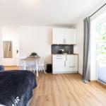 Schlafzimmer mit kleiner Küche und Essplatz. Holzboden, große Fenster und Zimmerpflanze.