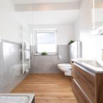 Badezimmer mit Dusche, Waschbecken und Toilette. Holzboden und graue Fliesen.