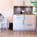 Küche mit Arbeitsfläche, Spüle und Kaffeemaschine. Nebenher Tisch mit Stühlen und Pflanze.