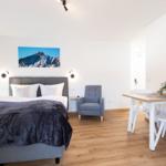 Schlafzimmer mit Bett, Sessel und Esstisch. Wandbild zeigt Berglandschaft.