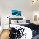 Schlafzimmer mit Bett, Wandbild und Sitzgelegenheit. Holzboden und Fenster mit Vorhang.