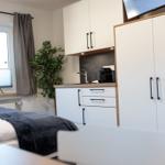 Schlafzimmer mit kleiner Küche und TV. Fenster mit Vorhängen und Zimmerpflanze.