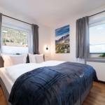 Schlafzimmer mit Doppelbett, zwei Fenstern und Wandbild. Holzboden und graue Vorhänge.