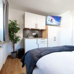 Schlafzimmer mit Bett, Kleiderschrank und Küchenzeile. Fenster mit Vorhängen und Zimmerpflanze.