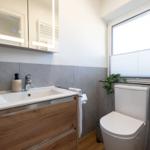 Badezimmer mit Waschbecken, Spiegel und Toilette. Fenster mit Jalousie und Pflanze.