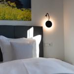 Schlafzimmer mit Bett, Kopfkissen und Wandlampe. Wandbild zeigt gelbe Blumen.