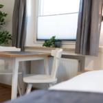 Ein Bett mit grauem Kopfteil, ein weißer Tisch mit zwei Stühlen und eine Fensterbank mit Pflanzen.