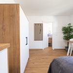 Ein Raum mit Holzboden, Bett, Tisch, Stühlen und Pflanze. Tür führt ins Bad.