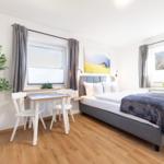 Ein Schlafzimmer mit Bett, Tisch und Stühlen, Fenstern und Zimmerpflanze.