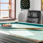 Billiard table with balls and cues. Dartboard and leather chair in background.