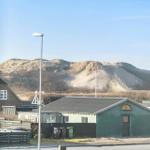 From a window, you see a green cabin, a road, and dunes with a flag.