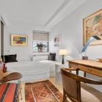 Bedroom with bed, desk, and wall art. Wooden chairs and rug on light walls.