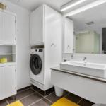 Bathroom with washing machine, mirror, and sink. Yellow rugs and white cabinets.