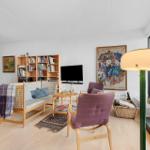 Living room with sofa, chairs, bookshelf, and TV. Large windows and artwork on the walls.
