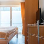 Schlafzimmer mit Bett, TV und Balkontür mit orangefarbenen Vorhängen.