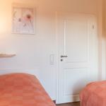 Zwei Betten mit orangefarbenen Bettdecken, weiße Tür und Wandregale mit Bildern.