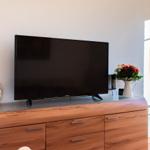 TV auf Holzboard mit Blumen und Dekoration. Fenster mit Vorhängen rechts.