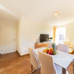 Essbereich mit Tisch, Stühlen und Obst. Wohnzimmer mit Sofa und TV. Küche mit Arbeitsplatte.