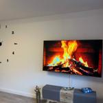 Wanduhr mit Ziffern an der Wand, Fernseher zeigt Kaminfeuer.