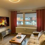 Wohnzimmer mit Sofa, Couchtisch und Fernseher an der Wand. Fenster mit Vorhängen und Heizkörper.