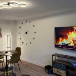 Wohn- und Essbereich mit Wanduhr, Fernseher und Tisch mit Stühlen.