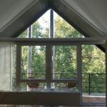 Large window overlooking balcony and forest. Wooden floor and gabled roof.