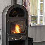 A wood-burning stove with a fire is positioned in front of a window with curtains.