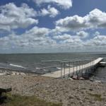Wooden pier leads to the water. Rocky ground and sky with clouds.