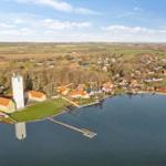 Aerial view of a village by water with houses, a church tower, and surrounding fields.
