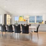 Dining room with table, chairs, and kitchen area. Large windows with view of terrace.