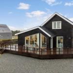 Modern wooden house with large balcony and glass front. A gray building is in the background.