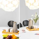 Round table with ceramic set, fruit, and flowers under a ceiling lamp.