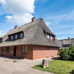 Haus mit strohgedecktem Dach, roten Ziegelmauern und grüner Umgebung unter blauem Himmel.