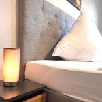 Ein Bett mit grauem Kopfteil und weißer Bettwäsche. Eine Lampe auf einem Nachttisch.