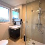 Badezimmer mit Dusche, Waschbecken und Fenster. Toilette und Spiegelschrank sichtbar.