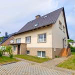 Ein zweistöckiges Haus mit braunem Dach und hellen Wänden. Eingangstreppe und gepflasterte Zufahrt.