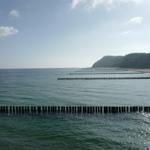 Strand mit Holzpfählen im Wasser und bewaldetem Hügel im Hintergrund.