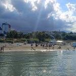 Strand mit Steg, Badegästen und Hotels im Hintergrund unter bewölktem Himmel.